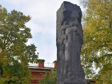 Karl Marx Monument on a tiered stone base with Cyrillic inscriptions, set among trees and a red brick building.