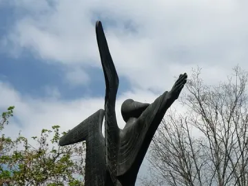 Angel of Peace sculpture with raised arms against a cloudy sky, standing among trees near a mural-covered building.