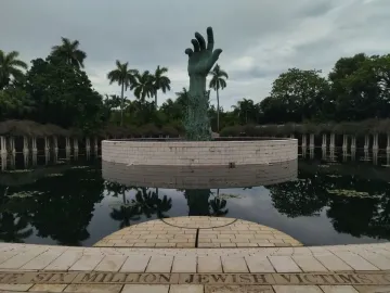 The Holocaust Memorial sculpture in Miami Beach shows a verdigris arm and hand rising from a circular stone base in a reflecting pool, with palm trees in the background.