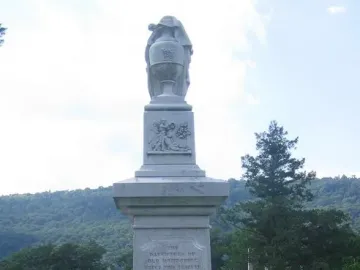 Confederate Memorial stone monument with a draped urn and carved relief in a cemetery, with a small Confederate flag and flowers at its base.