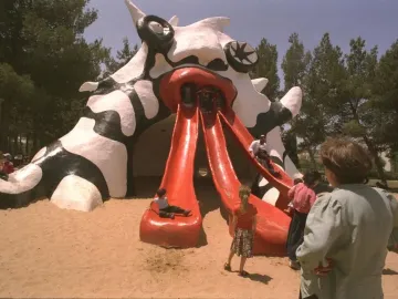 The Golem monster slide sculpture with three red chutes, with children playing in a sandy park.