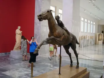 The Jockey of Artemision bronze sculpture displayed in a museum, showing a rearing horse with a small rider leaning forward.