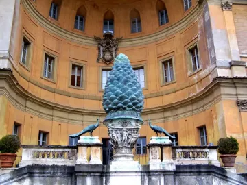 La Pigna pine cone sculpture on an ornate pedestal, flanked by two peacock statues in front of a curved orange building.