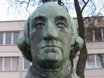 Weathered green-patinated bust of George Washington on a speckled stone base, with a building and bare branches behind it.