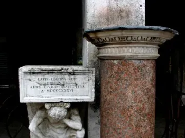 Il Gobbo di Rialto, a white marble crouching figure holding up an inscribed plaque beside a reddish-brown column.