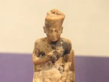 Khufu statuette of a seated ivory figure with a tall crown, shown on a clear base.