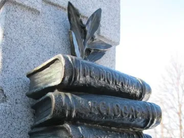 La mort de Dante monument detail showing four stacked dark metal books mounted on a stone pillar, with leafy decoration above the top volume.