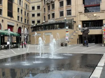 Nelson Mandela statue with raised arms in a busy public square, standing behind a fountain.