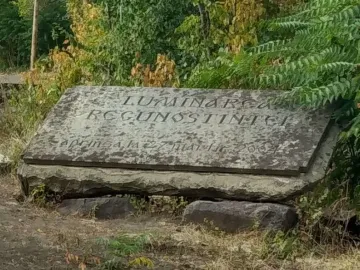 Weathered stone Thanksgiving Candle monument with a carved inscription on a tilted rectangular slab, set among rough stones and greenery.