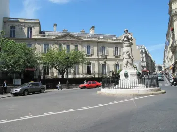 The Gavarni Fountain stands on a street corner behind a black metal fence, with sculpted figures, parked cars, and nearby buildings under a blue sky.