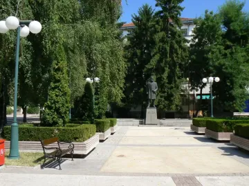 Monument to Hristo Botev: a bronze-colored statue of a bearded man holding a rolled document on a tall stone pedestal among trees.