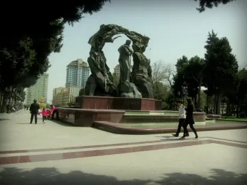 Huseyn Javid Monument with a central standing figure and two arcing bronze figures over a red base beside a fountain in a park.