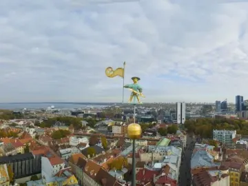 Old Thomas weathervane above Tallinn’s skyline with historic rooftops and modern buildings in the distance.