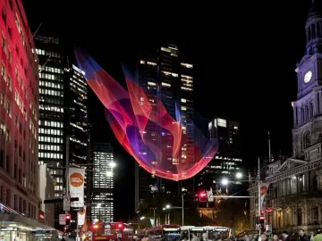 A large pink, red, and blue mesh sculpture hangs above a busy Sydney intersection at night with pedestrians and city traffic below.