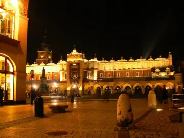 Adam Mickiewicz Monument lit at night in a city square with an arcaded building and clock tower behind it.
