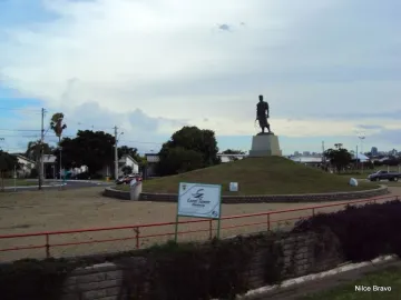 Laçador statue on a pedestal in a traffic circle, with houses, trees, and a cloudy sky behind it.