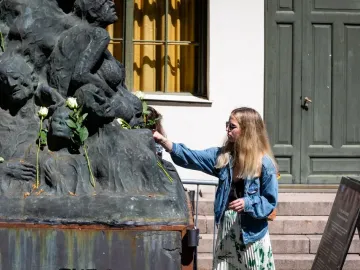 Pillar of Shame sculpture with white roses as a woman places another rose on it