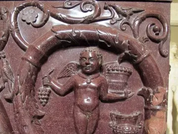 Red porphyry relief on the Sarcophagus of Constantina with a winged putto holding grapes beside a basket, framed by an elaborate vine garland.