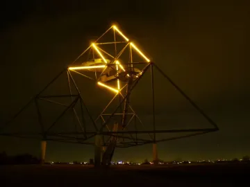The Tetrahedron sculpture lit at night with yellow lights along its upper edges and a spiral staircase inside.
