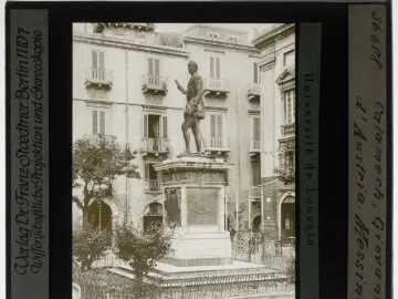 John of Austria monument on a tall ornate pedestal in a public square, with buildings and balconies in the background.