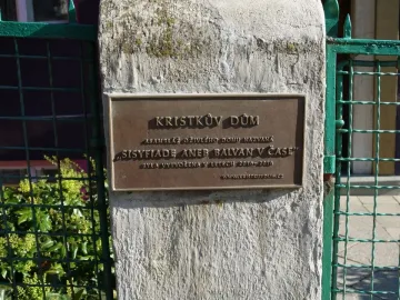 Bronze plaque for Kristek House mounted on a light-colored pillar beside a green metal fence.