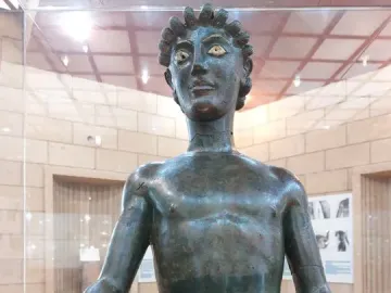 Efebo de Selinunte, a dark bronze nude young man statue with one arm raised in a museum display.