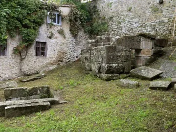 Etruscan tombs made of grey stone in a grassy outdoor area, with a vine-covered building and stone wall nearby.
