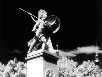 Athena Protects the Young Hero statue on a tall pedestal beside a sidewalk with people walking below.