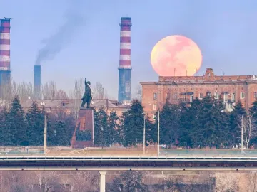 Lenin Monument in Zaporizhzhia with a raised arm, set on a tall pedestal near trees, industrial chimneys, and a large red moon behind a building.