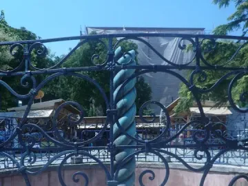 A section of the Serpent Column beside an ornate black wrought-iron fence, with a stone plaque block in the foreground.