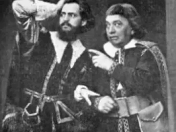 Two actors in elaborate stage costumes from The Stone Guest, one bearded man holding a hand to his head while the other points with both hands.