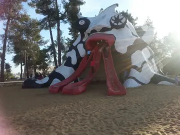 The Golem sculpture with children climbing its three red tongue-like slides in a sandy playground surrounded by trees.