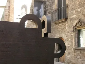 Topos V, a dark geometric sculpture with cutouts, stands in front of historic stone buildings in Barcelona.