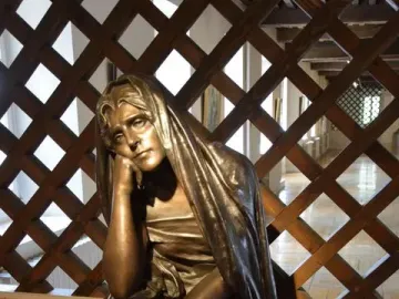 La Douleur bronze sculpture of a seated figure with its head resting on one hand before a wooden lattice screen.