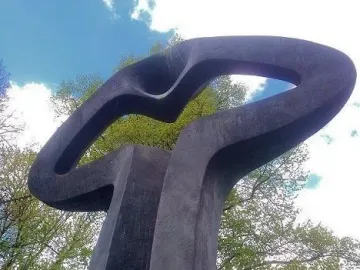 Der Mahner, a dark abstract sculpture with an open loop at the top, framed by green trees against a blue sky.