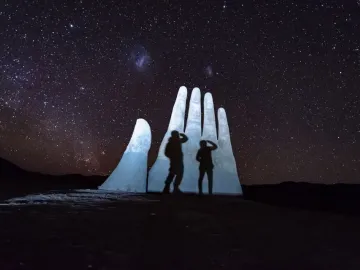 Mano del Desierto sculpture lit against a star-filled night sky, with two people standing in front of it.