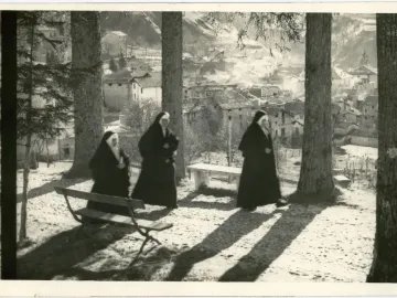 Three nuns walking along a tree-lined path with a village on the hillside in the background.