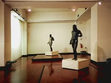 The Riace bronzes displayed on plinths in a museum gallery, with one larger bearded male statue beside a smaller figure.