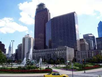 Swann Memorial Fountain spraying high jets in a plaza with skyscrapers and landscaped flower beds nearby.