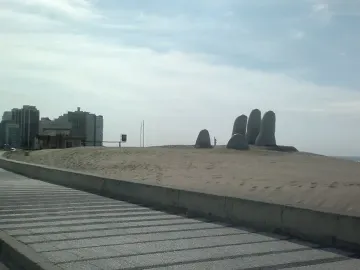 The Hand sculpture with five rounded fingers emerging from a sand dune, with a walkway, cityscape, and ocean in the background.