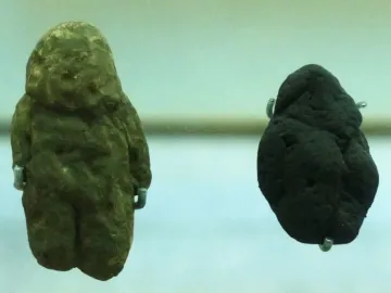 Two rough stone replicas of the Venus of Berekhat Ram, one greenish-brown and figure-like, the other darker and more irregular.