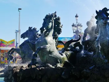 Bronze fountain at the Monument aux Girondins with rearing horses and spraying water, with a fairground attraction and ornate lamppost in the background.