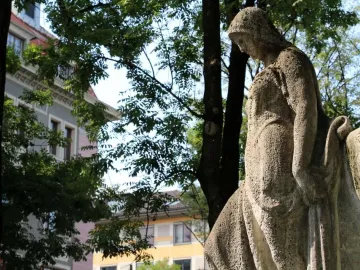 Weathered stone figure from the Nornenbrunnen stands beside trees and colorful buildings in the background.
