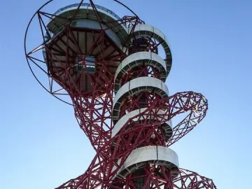 Orbit Tower’s red metal framework with a white spiral walkway rising to the top against a blue sky.