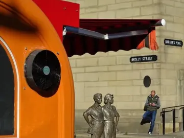 The Women of Steel sculpture stands outdoors on a paved area near a stone building, with street signs and a person in the background.