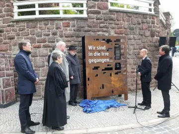 Rusted Dejudaization Institute memorial with German text and abstract cut-outs, surrounded by several people during an outdoor unveiling.