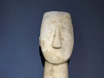 Kykladic Idol marble figurine shown from the upper torso up, with a long neck and stylized head against a dark background.