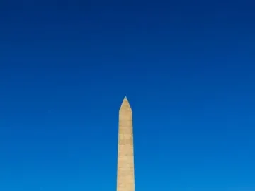 National Women\'s Monument obelisk with a bronze woman and child sculpture at the base, framed by trees and shrubs.