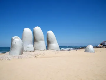 The Hand sculpture rising from the sand on a beach with the ocean and blue sky in the background.