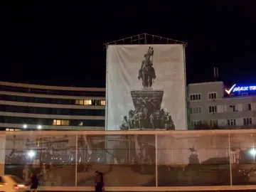 Tsar Liberator Monument covered by a large white banner on scaffolding at night, with nearby buildings and street activity.